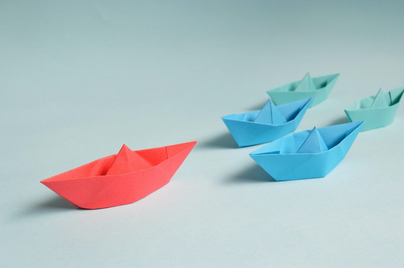 A red paper boat heading a different way from the blue boats